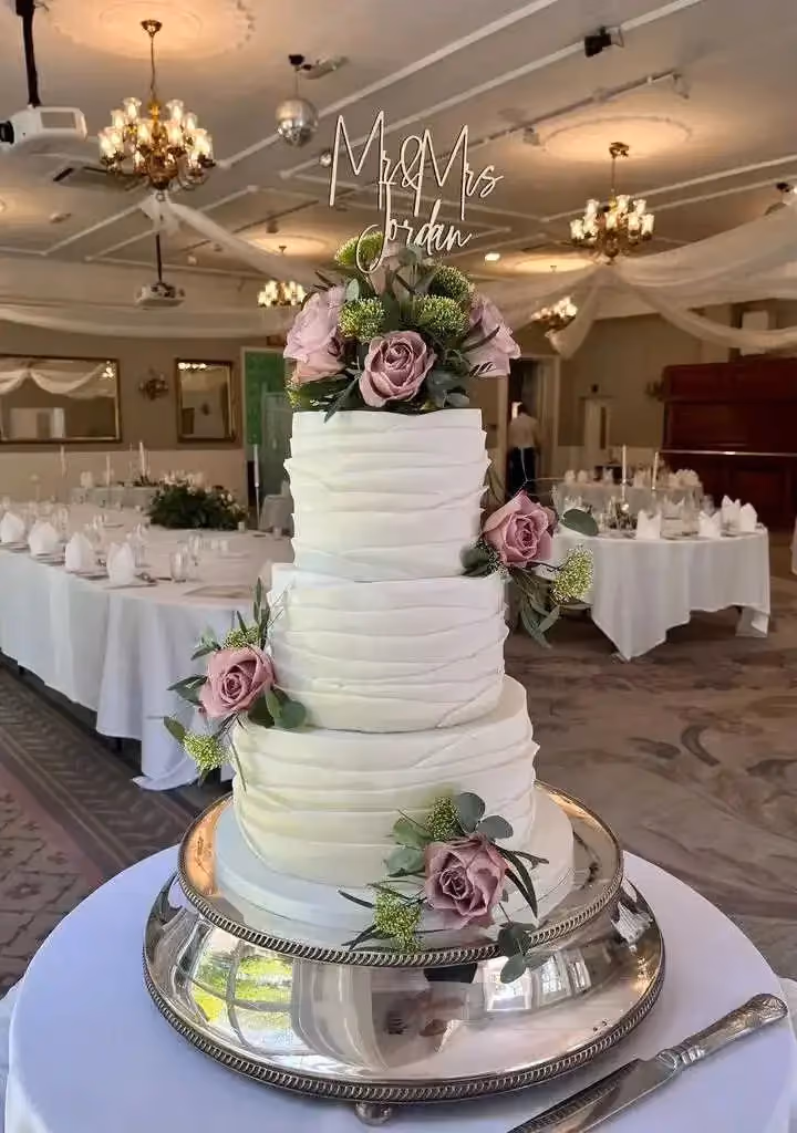 A three-tier white wedding cake with textured frosting, decorated with dusty pink roses and green foliage, topped with a 'Mr & Mrs Jordan' sign. The cake is on a silver stand in a wedding reception room.