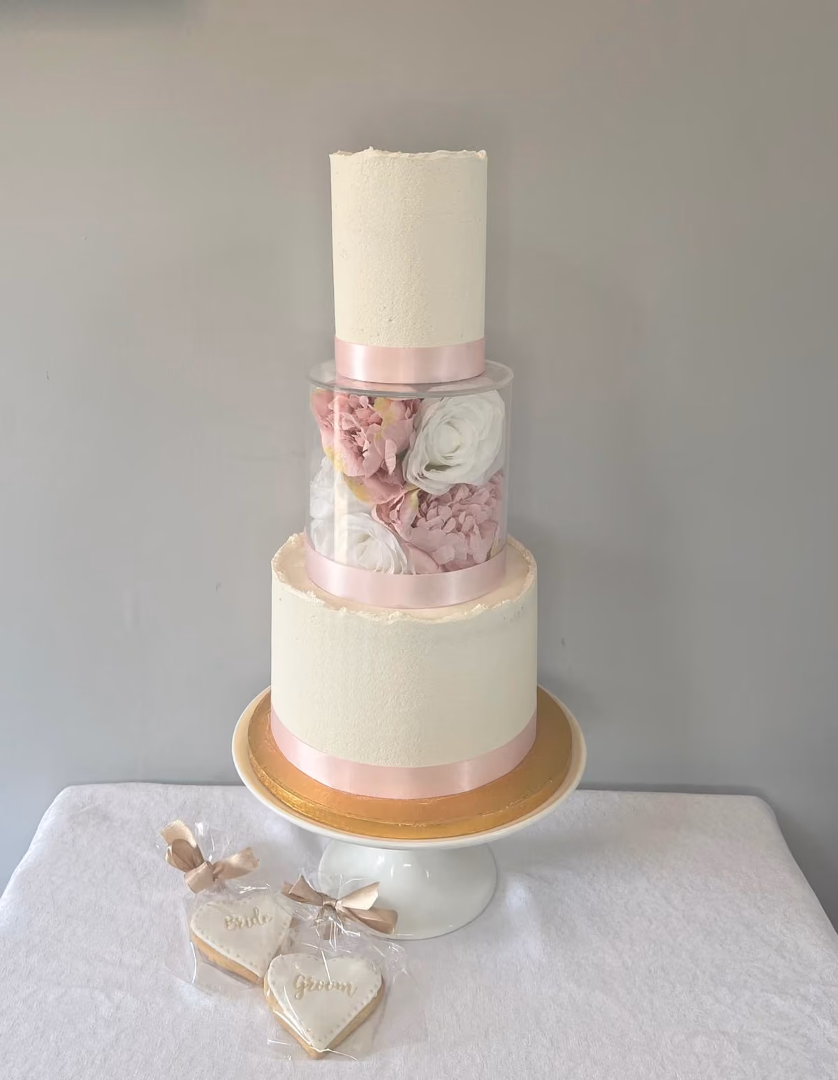 A tall two-tier ivory buttercream wedding cake with raw-edged tops and pale pink satin ribbons, separated by a clear acrylic floating tier filled with pink peonies and white roses. It sits on a white cake stand with a gold board, beside wrapped heart-shaped 'Bride' and 'Groom' biscuits.