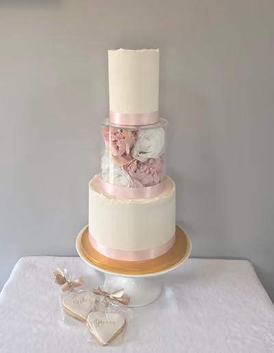 A tall two-tier ivory buttercream wedding cake with raw-edged tops and pale pink satin ribbons, separated by a clear acrylic floating tier filled with pink peonies and white roses. It sits on a white cake stand with a gold board, beside wrapped heart-shaped 'Bride' and 'Groom' biscuits.