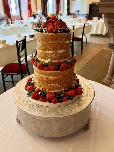 A two-tier naked wedding cake with visible sponge layers and cream filling, dusted with icing sugar and topped and encircled with fresh strawberries, raspberries, blueberries and small white flowers. It sits on an ornate silver cake stand in a wood-floored reception room.