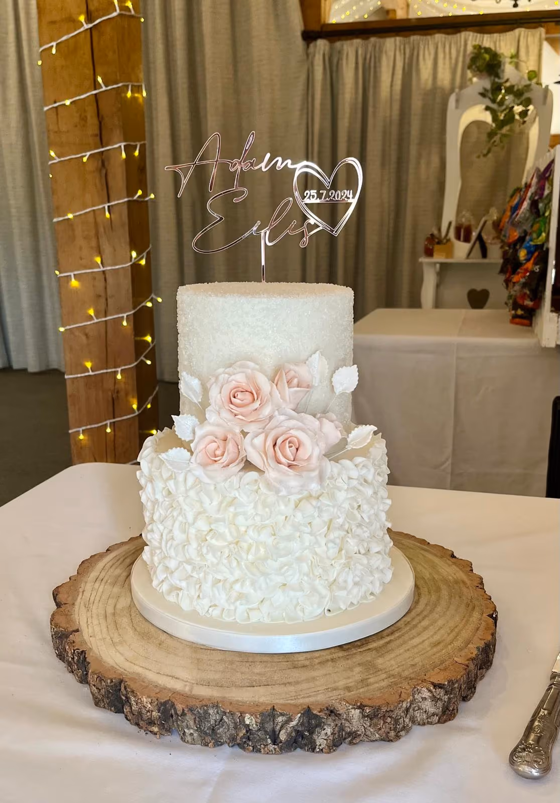 A two-tier wedding cake with a sparkling white top tier and a ruffled white bottom tier, adorned with pink sugar roses and white leaves. A rose gold cake topper features the names Adam and Eilís with the date 25.7.2024, and the cake sits on a rustic wooden log slice.