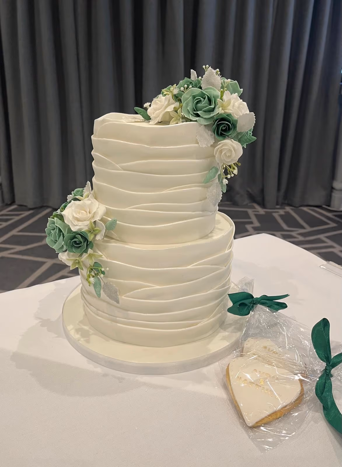 A two-tier white wedding cake with a ruffled fondant design, decorated with clusters of white and green sugar flowers and greenery. Beside it is a heart-shaped cookie in clear packaging tied with a green ribbon.
