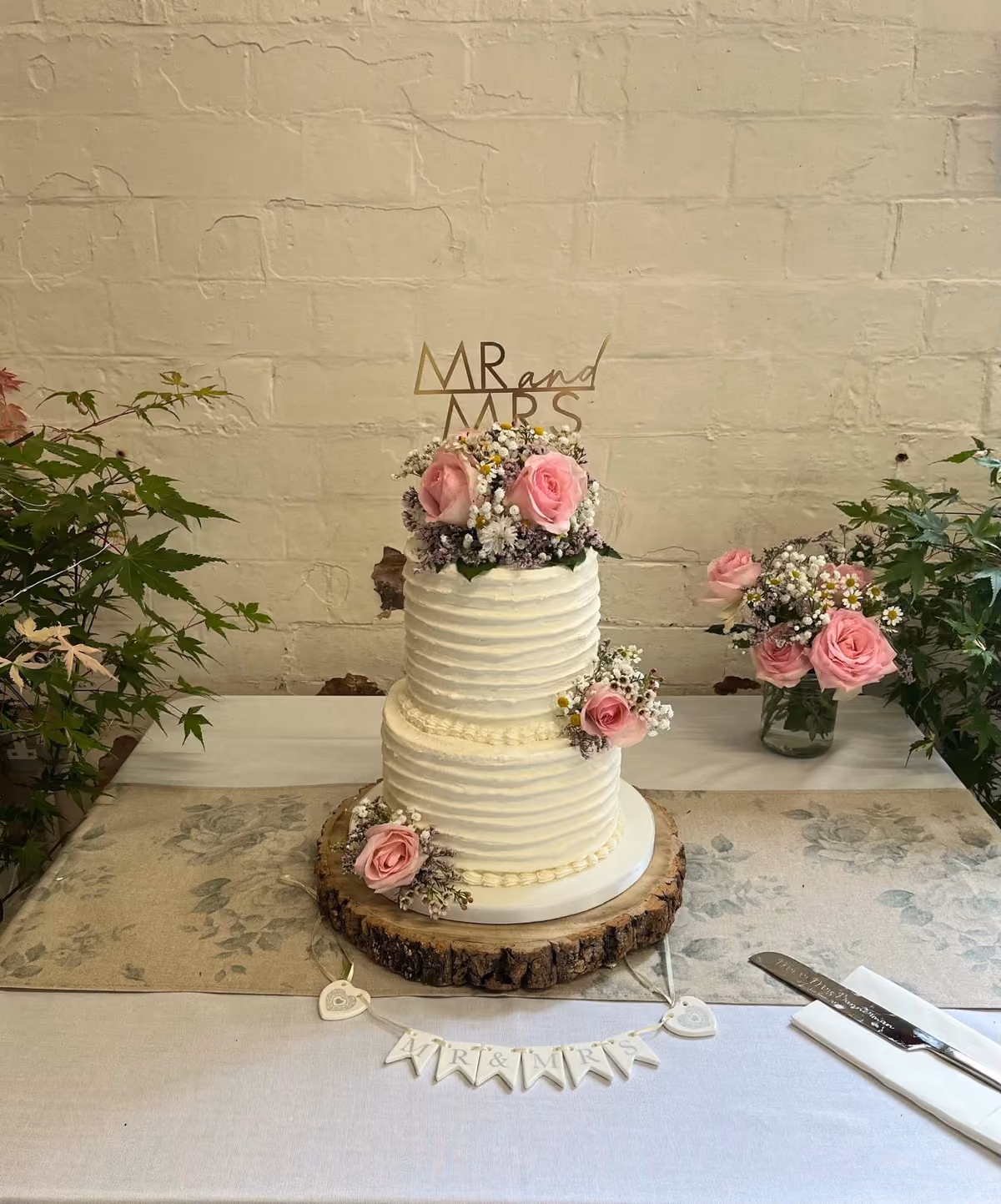 A two-tier wedding cake with textured white buttercream, decorated with pink roses and small white and purple flowers, topped with a gold 'MR and MRS' topper, sitting on a rustic wooden stand.
