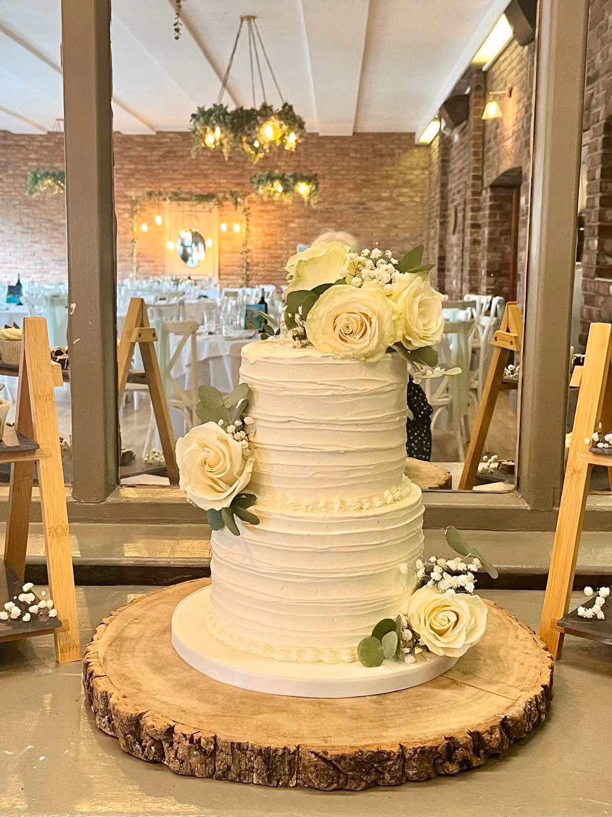 A two-tier white wedding cake with a textured buttercream finish and piped borders, decorated with fresh white roses, eucalyptus and baby's breath. It stands on a wood slice in a brick-walled venue with festoon lights and greenery chandeliers.