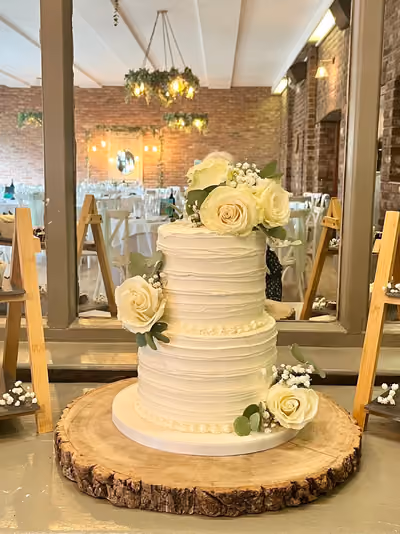 A two-tier white wedding cake with a textured buttercream finish and piped borders, decorated with fresh white roses, eucalyptus and baby's breath. It stands on a wood slice in a brick-walled venue with festoon lights and greenery chandeliers.