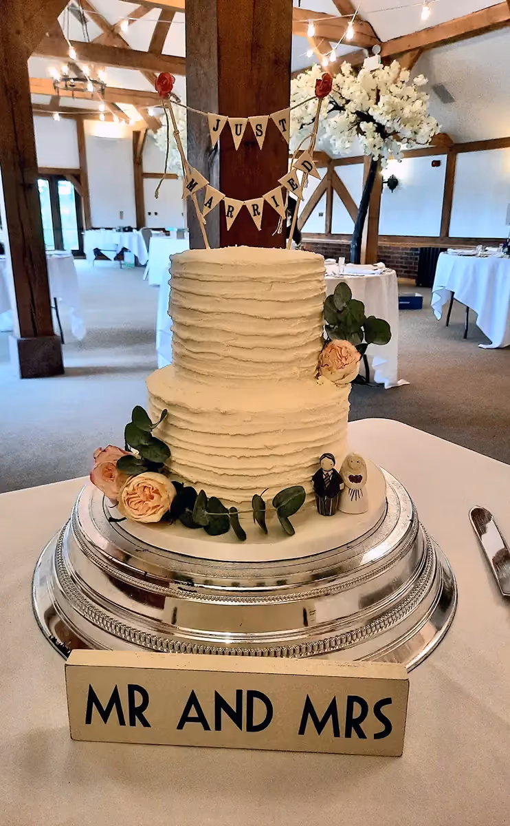 A two-tier wedding cake with textured cream frosting, decorated with peach roses, eucalyptus leaves, a 'Just Married' banner cake topper, and small bride and groom figurines. A 'MR AND MRS' sign sits in front of the cake.