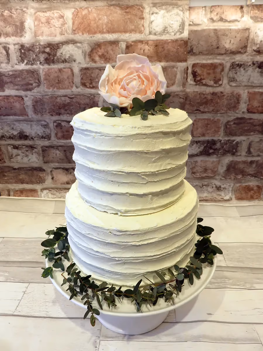 A two-tier wedding cake with white textured buttercream, topped with a large light pink sugar flower and green leaves, and decorated with eucalyptus around the base.