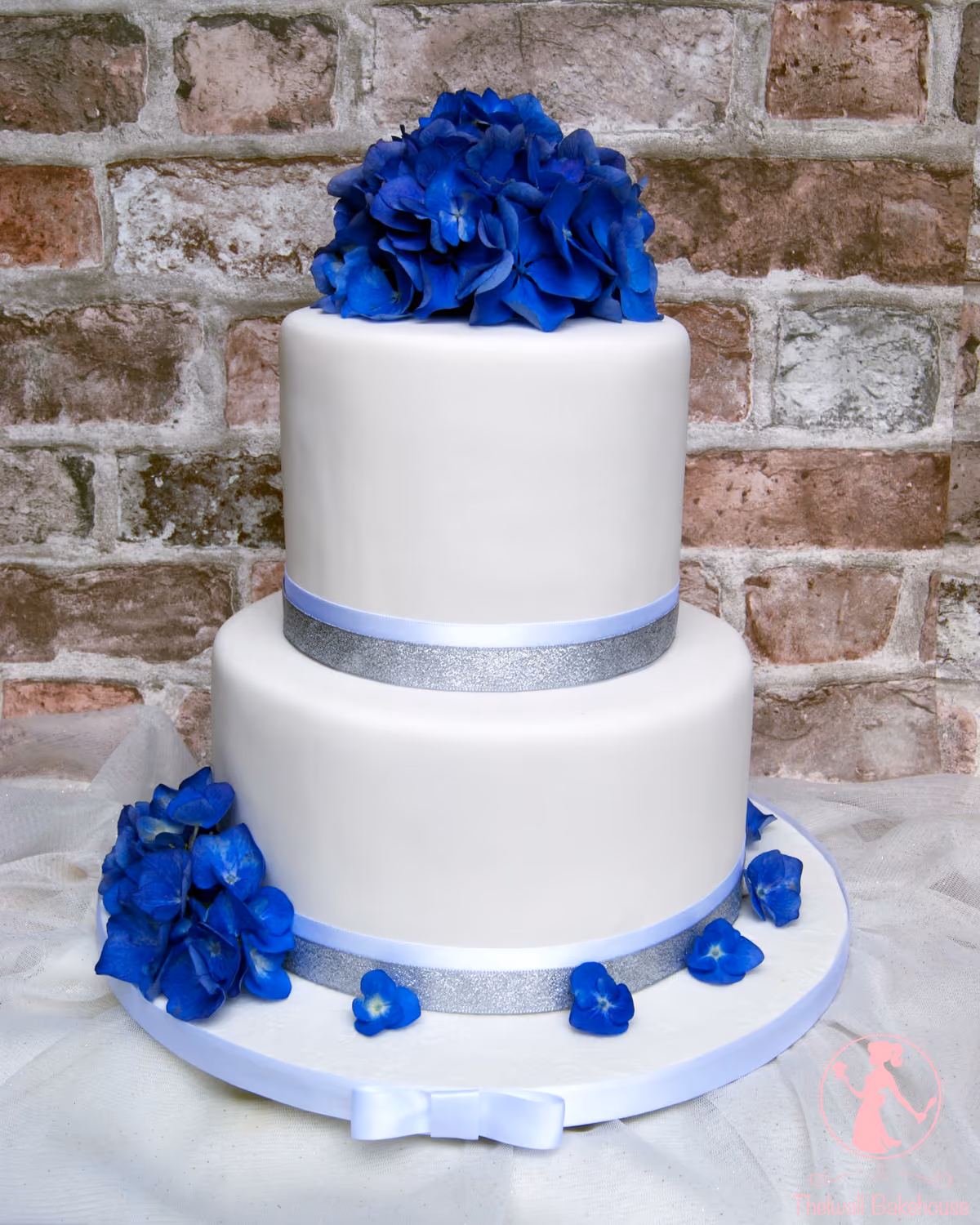 A two-tier white fondant wedding cake decorated with vibrant blue hydrangeas on top and a cluster on the lower tier, accented with light blue and silver glitter ribbons and a small white bow on the cake board.