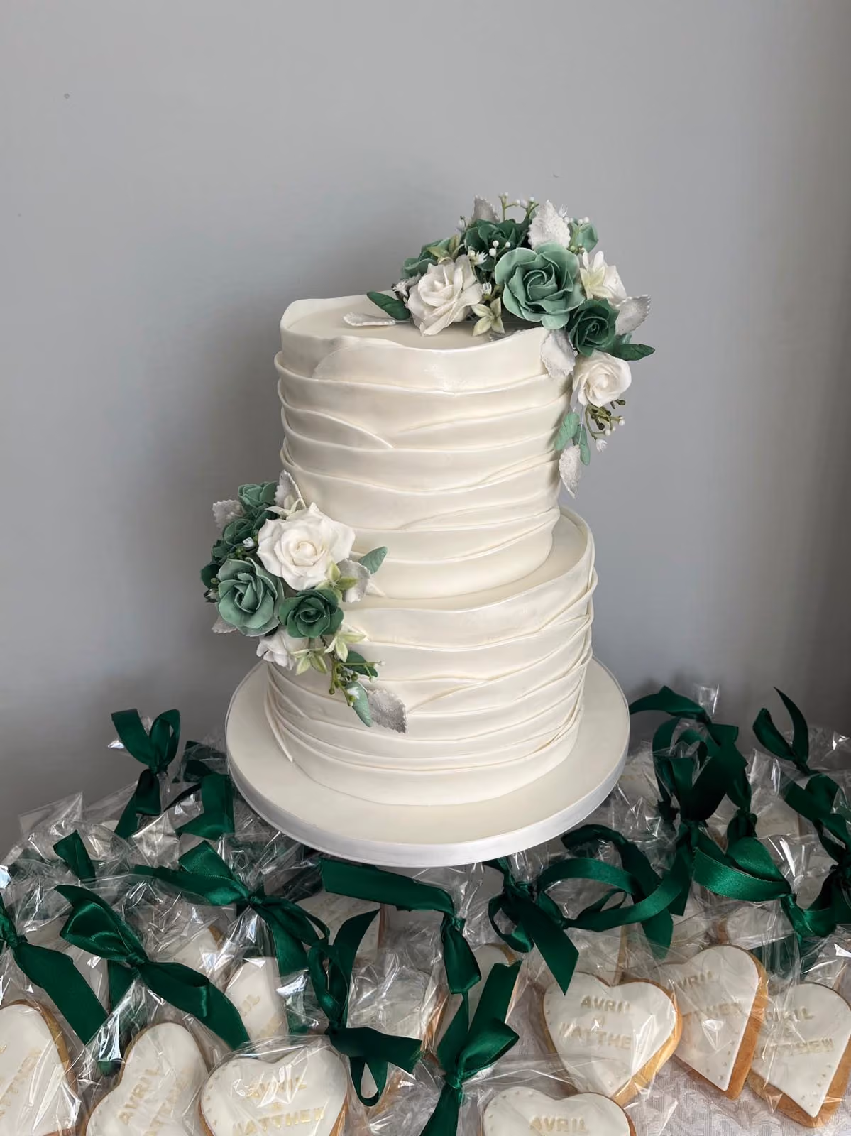 A two-tier white wedding cake with layered, ruffled fondant wraps, decorated with clusters of green and white sugar roses and silver leaves. It is surrounded by heart-shaped favour biscuits reading 'Avril & Matthew', individually wrapped and tied with green satin ribbons.