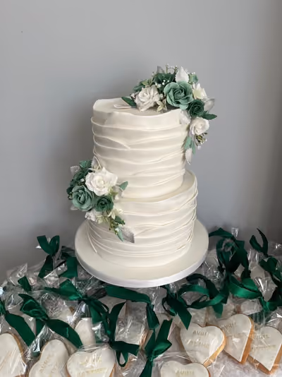 A two-tier white wedding cake with layered, ruffled fondant wraps, decorated with clusters of green and white sugar roses and silver leaves. It is surrounded by heart-shaped favour biscuits reading 'Avril & Matthew', individually wrapped and tied with green satin ribbons.