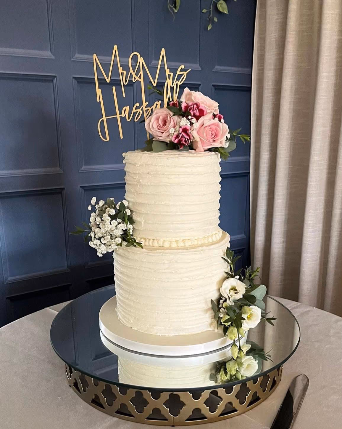 A two-tier white wedding cake with textured buttercream frosting, decorated with pink roses and white baby's breath flowers, and topped with a gold 'Mr & Mrs Hassall' cake topper. The cake sits on a mirrored stand.