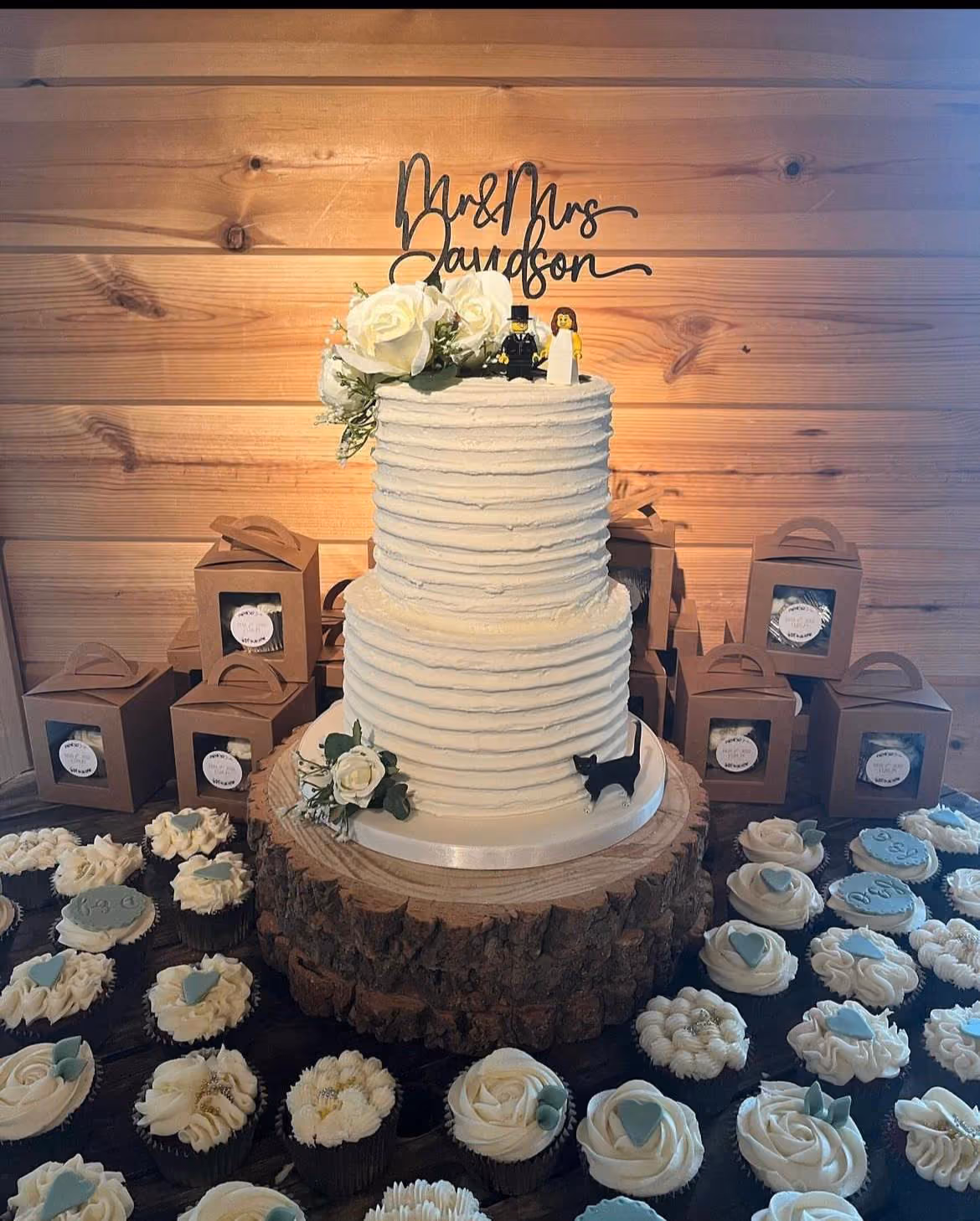 A two-tier white wedding cake with textured frosting, topped with a 'Mr & Mrs Davidson' sign, white roses, and Lego bride and groom figures. A black cat silhouette decorates the base, which rests on a rustic log stand. Many white and light blue frosted cupcakes surround the cake, set against a wooden wall and gift boxes.