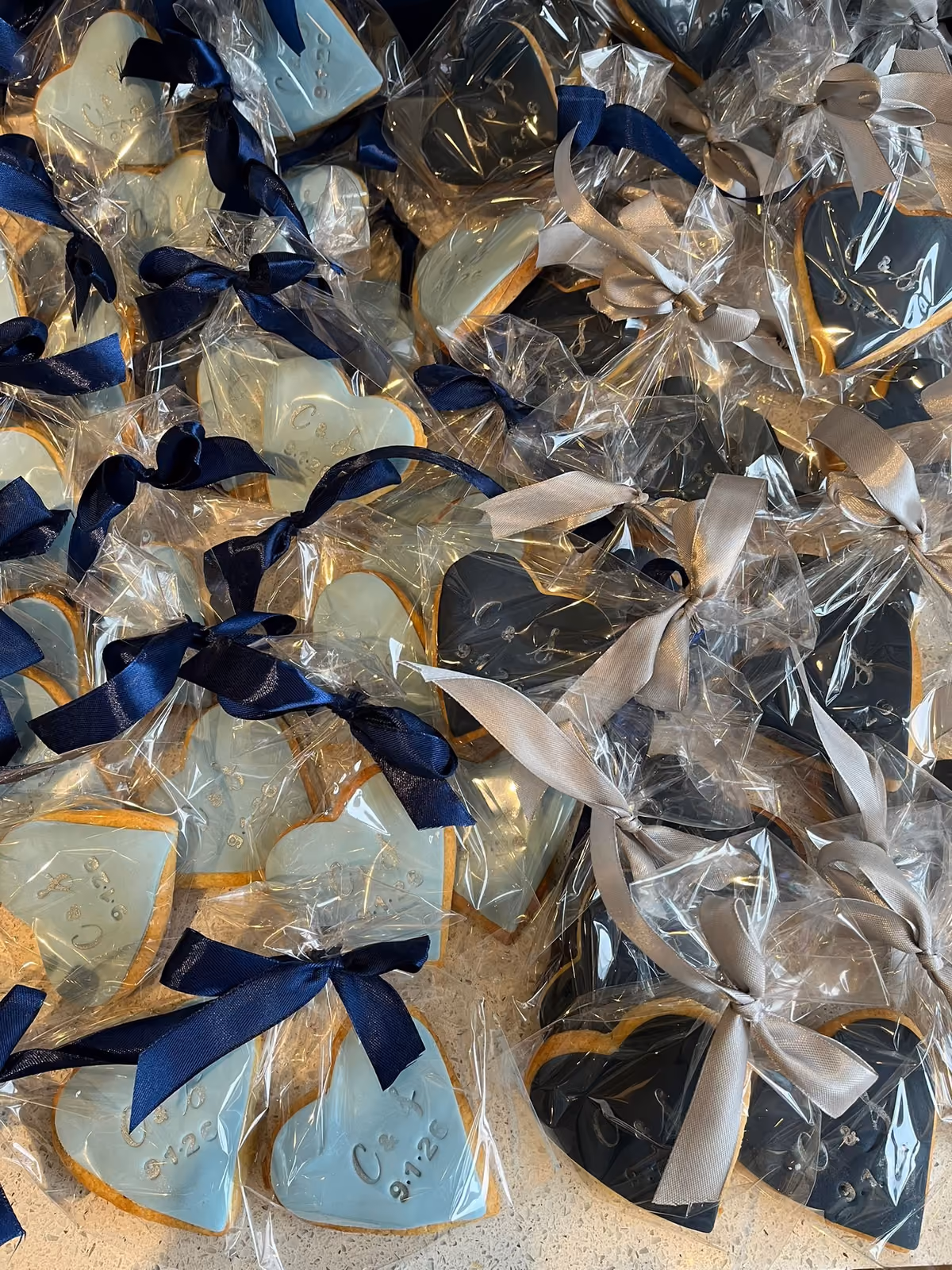 Numerous heart-shaped wedding favor biscuits, individually wrapped in clear plastic and tied with ribbons. Some biscuits are light blue with navy ribbons, and others are dark grey with silver ribbons, each embossed with 'C & J' and '9.7.26'.