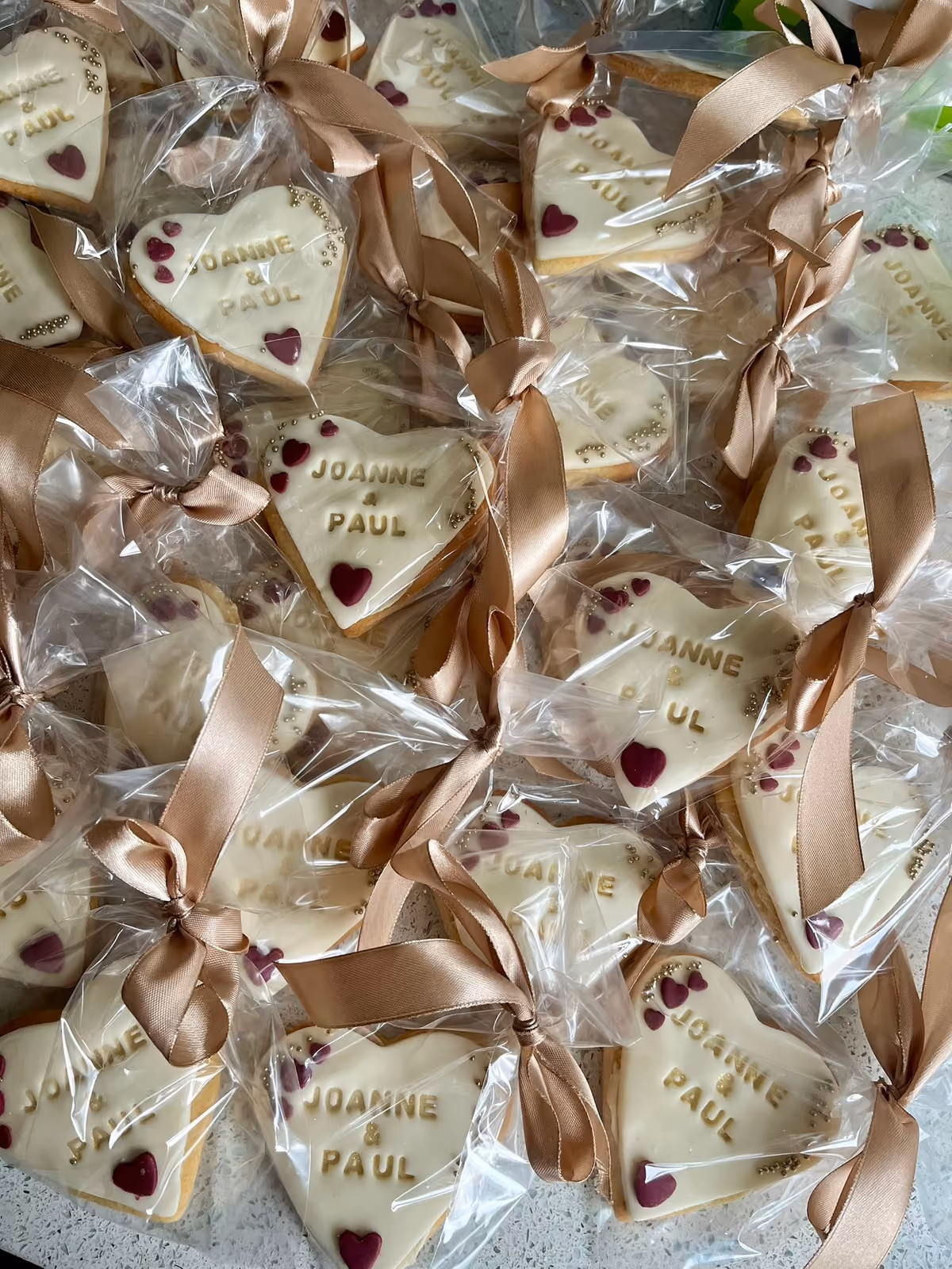 Numerous individually wrapped heart-shaped biscuits, decorated with white icing, gold lettering spelling 'JOANNE & PAUL', small burgundy hearts, and gold edible beads, each tied with a champagne-colored ribbon, arranged as wedding favors.