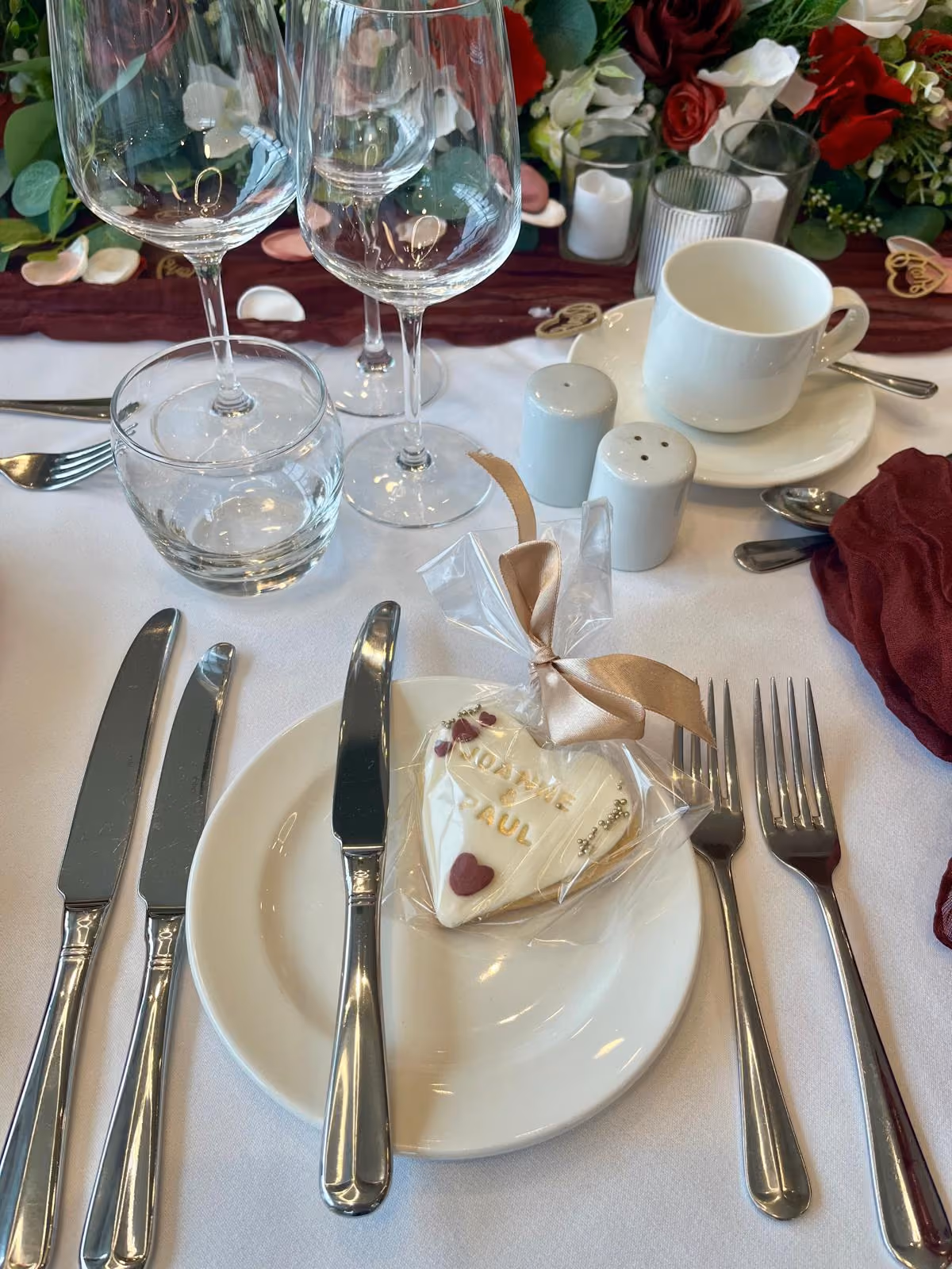A heart-shaped white iced biscuit with 'JOANNE & PAUL' written in gold, decorated with small red hearts and silver beads, individually wrapped with a gold ribbon, placed on a white plate at a wedding table setting with cutlery, glasses, and floral decor.
