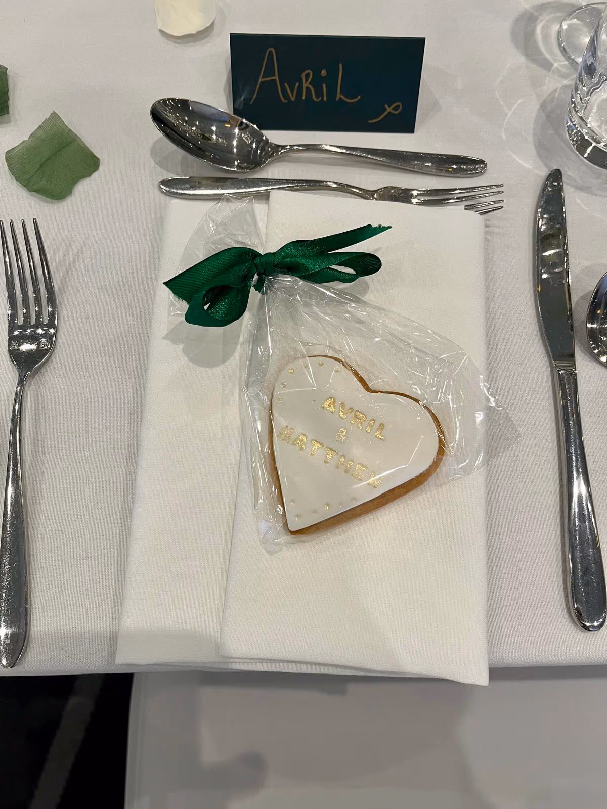A heart-shaped biscuit, iced in white with gold text 'Avril & Matthew', packaged as a wedding favour with a green ribbon, placed on a white napkin at a table setting.