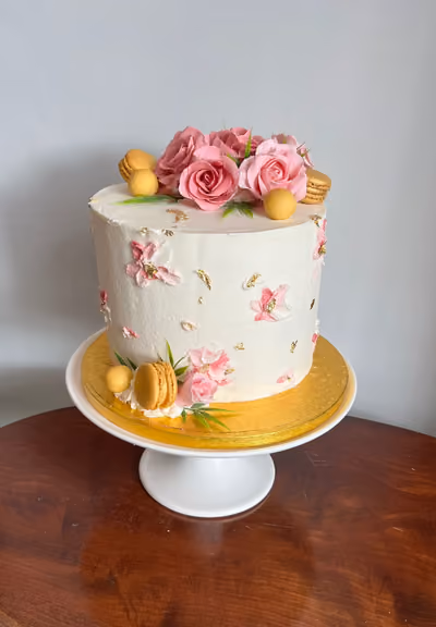 A single-tier white cake decorated with a cluster of pink sugar roses, gold macarons, and yellow spheres on top, and similar decorations with delicate pink buttercream flowers and gold leaf flakes around the sides. The cake is presented on a white cake stand with a gold base.