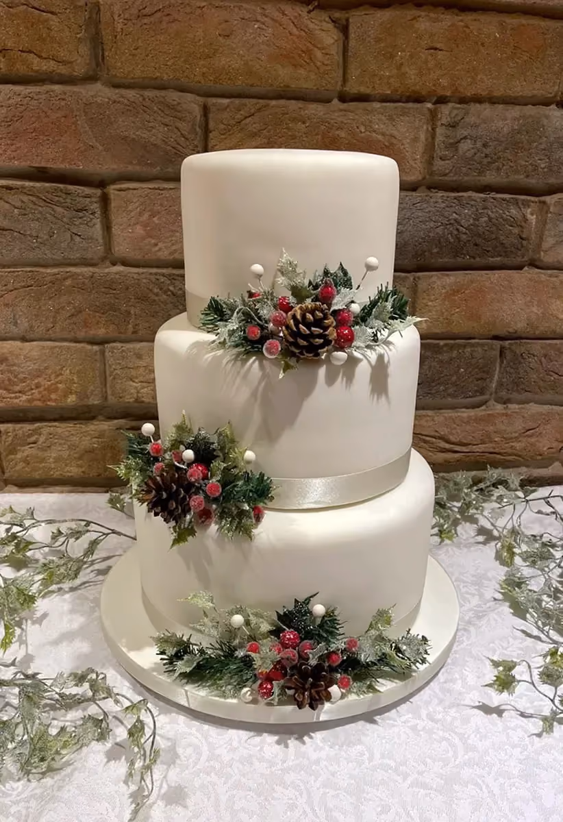 A three-tier white fondant cake decorated with frosted winter foliage, red berries, and pinecones, set against a rustic brick wall.