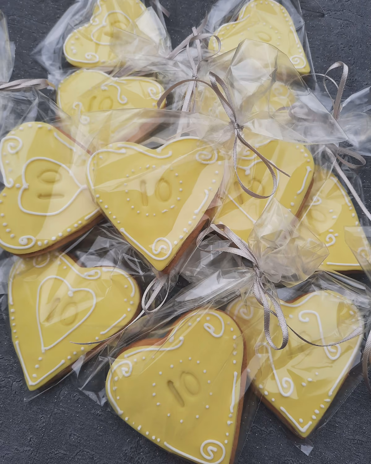Multiple yellow heart-shaped iced cookies, each decorated with white icing swirls, dots, and the number '10', individually wrapped in clear plastic with grey ribbons, arranged on a dark grey textured surface.